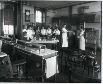 Class members cooking in a kitchen classroom.  'Lady standing at far left:  Miss Rachel Colwell; man in middle back row:  Thomas E. Hodges, President of WVU.'