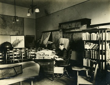 'Unidentified faculty member at his desk surrounded by maps and books. He is probably a member of the faculty of the Geography department, and his office is probably in Woodburn Hall.'