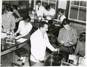 Chemistry students at University High School from the class of 1961. The students at the left table are Edwin Riffle and Janet Yaromy, those  at the right table are teacher Michael Caruso, Julius Mogroyos, and an unknown  student; those at the back table are Robert Weaver,an unknown student teacher, James Nicklow, and Julia Frazier.