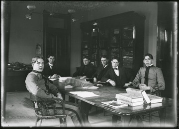 Six male students sitting around table with books, looking very serious.