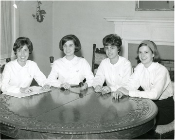 Female members of WVU Panhellenic officers gather around a round table in the office.  All four members wear white long sleeved shirts and dark pencil skirts.