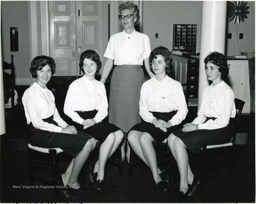 Female members of WVU Panhellenic officers who dressed uniformly in a white shirt and a dark skirt seated in the reception of Elizabeth Moore Hall for a photo shoot with Mary Jane Schuster 'now Mrs. English' standing in the middle. 