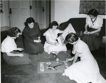 YWCA Members sit on the floor of the YWCA office and engage in making promotional posters for their spring cruise; Some of them are shown with leis around their neck; Betty Boyd is second from left.