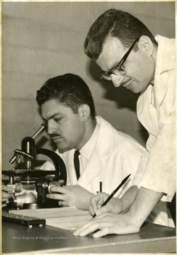 Two researchers in a lab coats are shown working; one seated looking through a microscope while others standing records it on a notebook.