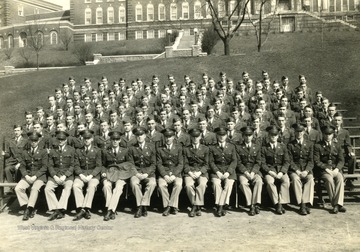 The cadets of company E sit on bleachers with officers for a group portrait on the Drill Field with Stalnaker Hall in the background. See original for identification.