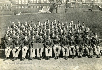 The cadets of company D sit on bleachers with officers for a group portrait on the Drill Field with Stalnaker Hall in the background. See original for identification.