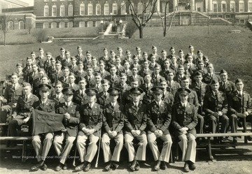 The cadets of company A sit on bleachers with officers for a group portrait on the Drill Field with Stalnaker Hall in the background. See original for identification.