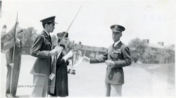 Sponsor holding trophy, cadet holding sword, commander reaching for trophy.