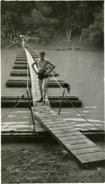 One cadet in a swimsuit nears the end of a pontoon bridge while others on the opposite bank watch.