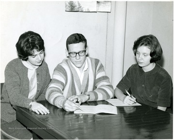Male student pointing to notebook with pen surrounded by two female students.