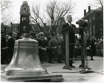 'Rev. Joe Gluck speaks at Dec. 7, 1967 dedication ceremonies for the bell from the armored cruiser 'U.S.S. West Virginia.'  Two other main speakers are shown seated in the first row: WVU President James B. Harlow (second from left) and Naval Reserve Captain Marlyn E. Lugar (third from left).