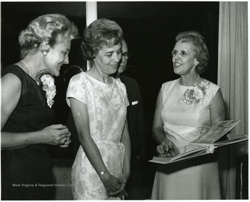 'Mrs. Gilbert Bachmann, member of the WVU Board of Governors (left), Mrs. Donovan H. Bond, wife of the exec. dir. of the 100th Ann. observance (center), and Mrs. Harry B. Heflin, wife of the acting president of WVU (right) look over a scrapbook presented to the Heflins.'