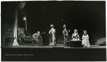 Women in Victorian clothing on stage next to a ornate carriage.