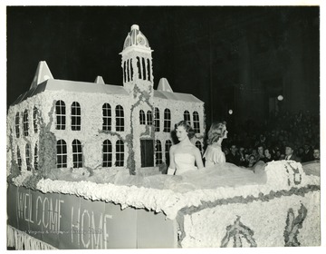 Students ride on a float of Woodburn Hall with a banner of 'Welcome Home' on the side.