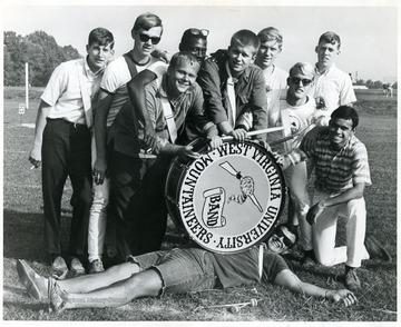 WVU band members pose around a bass drum: two students in darker shirt with drum sticks in hands and leaning over a drum are Theis brothers.