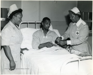 'Chief Nurse 1st Lt. Elizabeth T. Dozier, Rt. #1, Box 79, Eastover, South Carolina, looks on while Pfc. Calvin Williams, 1836 'E' Street, Fresno Calif., pleasantly passes away hours with handcraft in Lockbourne Army Base Station Hospital, Columbus, Ohio, with the aid of Franklin County Red Cross Chapter Gray Lady, Mrs. Lillie B. Williamson, 170 North Champion Avenue, Columbus, Ohio.'