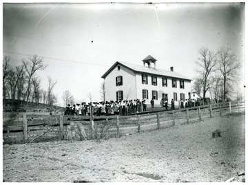 A view of Sinks Grove School House in Monroe County; many are outside on the school ground and some look out from the windows.