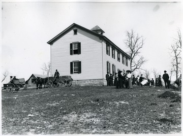 There are many people on the school ground; there are also a horse drawn buggies and a carriage.  This is possibly a school house in Sinks Grove in Monroe County.