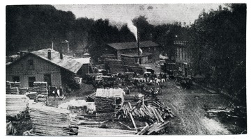 A view of a C&amp;O depot near Sandstone; there are several buildings, workers, horses and buggies, and piled lumber in sight.