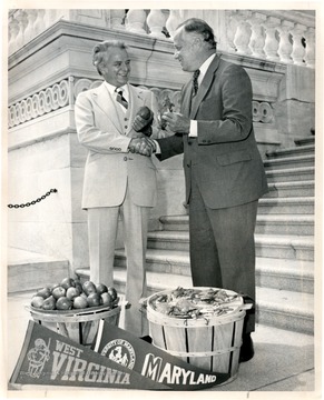 Robert C. Byrd shakes hands with another gentleman; barrels of fruit and W. Va. and Maryland pennants in foreground.