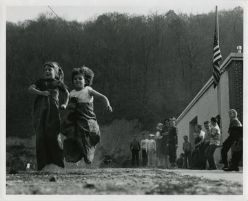 Two kids outside of a school sack racing while other students watch.