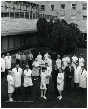 A group portrait of faculty and staff standing outside the WVU Medical Center. Included among the group are Clark Sleeth (front, third from left) and Eugene Stiles (center foreground, dark suit).