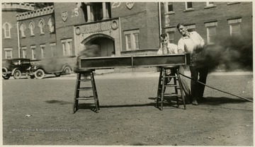 A photograph of two men working with a piece of equipment. WVU Armory visible in background.