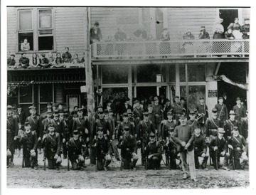 Unit of soldiers pose in the street wearing uniforms and holding weapons as civilians, including children sitting on the rooftop, watch.