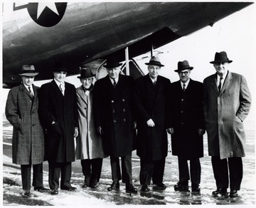 A group photograph of Congressmen gathered on the tarmac in front of an airplane.