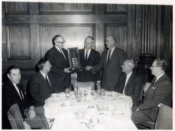 A photograph of Mr. Courtney (third from the left) being presented with a plaque. John Riley is seated on the far left.
