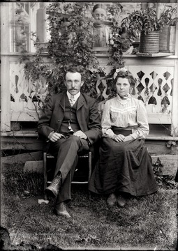 A portrait of seated man and young woman taken outdoor; while they sit their back against the porch, three girls on the porch peek through plants.  