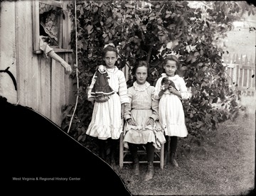 A portrait of three girls; one with a doll and the other with a puppy.  A woman stretches her arm out from the window toward them.