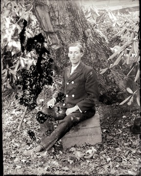 A portrait of man sitting on a wooden box near the tree.