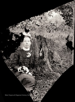 A portrait of young woman sitting by a tree stump; she is wearing a corsage at her shoulder and holding a posy.