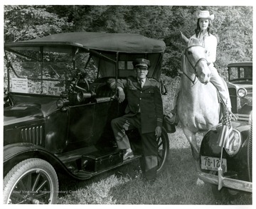 A man in a uniform leans against a 1920 Ford while a woman on horseback poses among several other antique automobiles. Both subjects are not identified.
