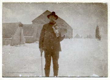 An unidentified male stands in the snow holding firewood in his arm.