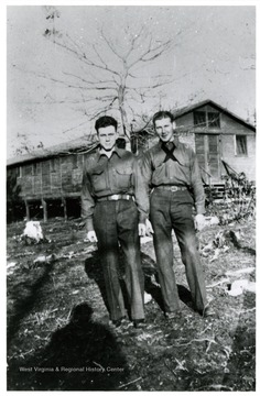 Two males stand, there is a barrack in the background.
