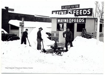 Three men shoveling snow in front of Wayne Feeds on the corner of School Avenue and Hewes Street near Kaiser Fraser Auto shop in Clarksburg.