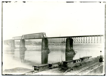 A truss bridge over the Ohio River; people sitting on a railway stretched along the river. 