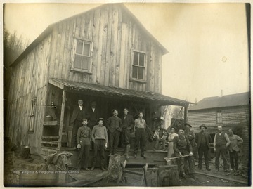 'This picture shows the family group of mostly sawmill men, part of whom lived with Mr. and Mrs. Martin L. Brown in the house shown and includes part of the office force and railroad crew. This group is made up as follows: Left to right: D. D. Brown(standing on the porch), Webb Helmick(standing next to D. D. B.), and Lester H. Sickler(engineer). Standing first on ground: Eugene Schock(sawyer), Charles W. Fenney(stenographer &amp; bookkeeper), Overton(handy-boy from Towanda, Pa.), Eliza Brown(Mrs. M. L. Brown), Fred Brown( with axe, locomotive engineer), W. W. Burton(Boer War, Englishman from England), J. B. White(saw filler), M. L. Brown(mill and yard superintendent) and Sag(setter).'