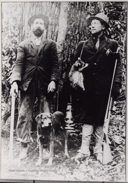Portrait of two hunters with a dog. Man on the right is holding a gun, a raccoon, and a lantern. The man on the left is holding an ax.
