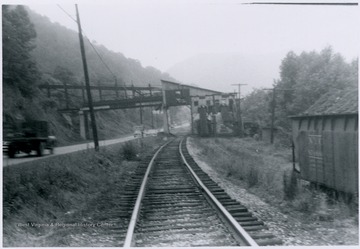 Portrait taken from the railroad tracks next to the highway. Cars and trucks can be seen passing underneath the conveyor. 