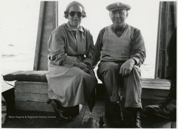 In this portrait, Menk and an unidentified woman are sitting together. Menk has on a hat and the woman is wearing sunglasses.