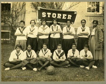 Team portrait of Storer College girl's basketball team in uniform in front of STORER banner.
