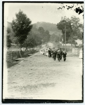 Several men and women, marching down a dirt road, many playing instruments.