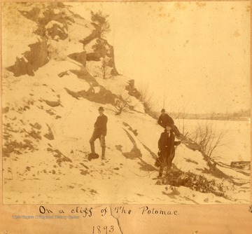 Three unidentified men wearing derbies and suits; carrying guns and dead animals, at the bottom of snow-covered cliffs.