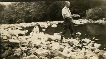 George Bird Evans fly fishing from a rocky bank while an unidentified woman watches.