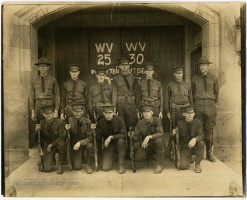 Group portrait of young men in uniform, probably West Virginia University students, posing in front of a university building. The doors of the building are labeled with "WV 25" and "WV 30".  Several students have Greek symbols written in ink on their left shoulder.