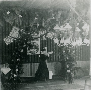 A woman positions a cross on the alter, which is decorated in evergreens and ornaments for Christmas. Note the kerosene lamp chandelier to the right. The church is located in the Clay District of Monongalia County.