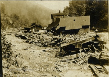 Postcard photograph of the flood destroyed buildings. One small house remains intact with a family standing outside and laundry hanging in the background. Information on the back: "Hinton Daily News Collection-John Faulkner Collection; from Jim Pettrey to Stephen Trail, 1997". See the back of the original for correspondence.